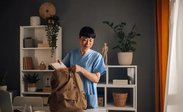 Asian dedicated nurse in blue scrubs is seen preparing medical tools and organizing documents into a brown backpack in a well-lit, modern room with shelves and assorted decor.